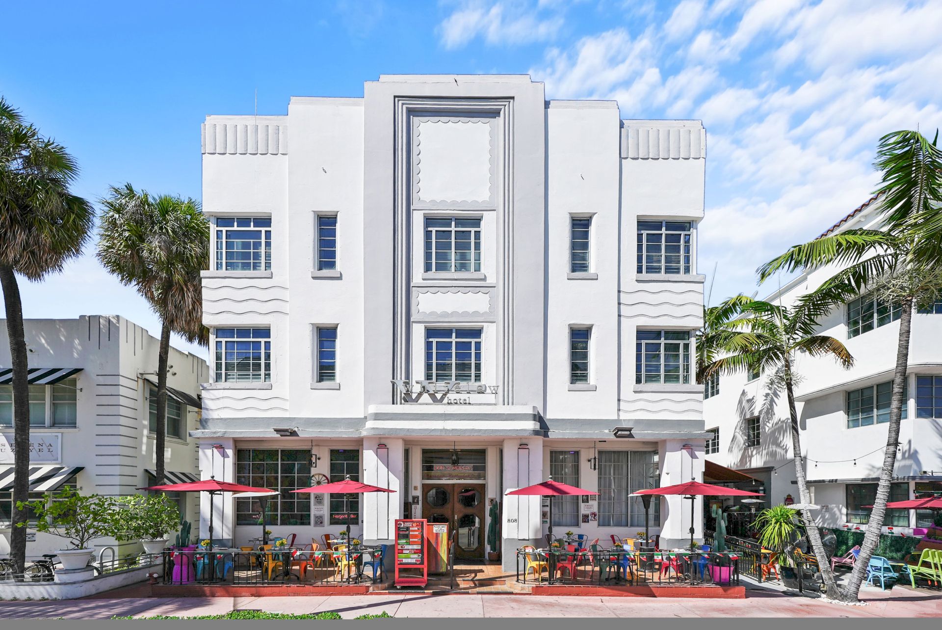 A stylish white Art Deco-style hotel with a central entrance, outdoor seating under red umbrellas, and palm trees lining the sidewalk.