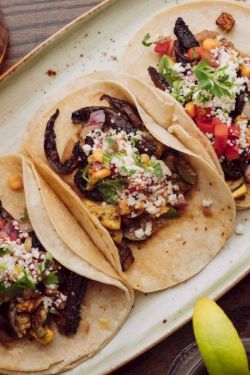 Three tacos topped with veggies and cheese on a plate, plus a lime wedge and drinks nearby, all on a wooden table.