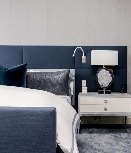 A modern hotel bedroom: blue bed with pillows, a nightstand with lamp, decorative piece, and a TV remote by the bed.