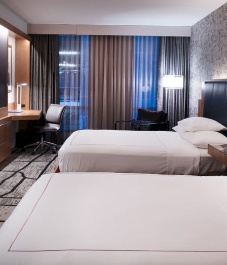 Modern hotel room with two beds, a TV, desk, and dark curtains by the window.