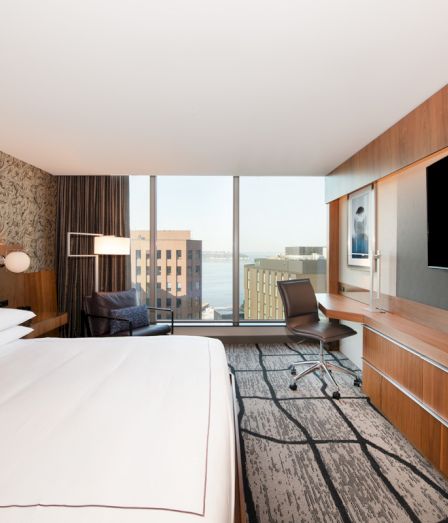 Modern hotel room with a large bed, desk chair, wall-mounted TVs, wood paneling, and big windows showing a city view.