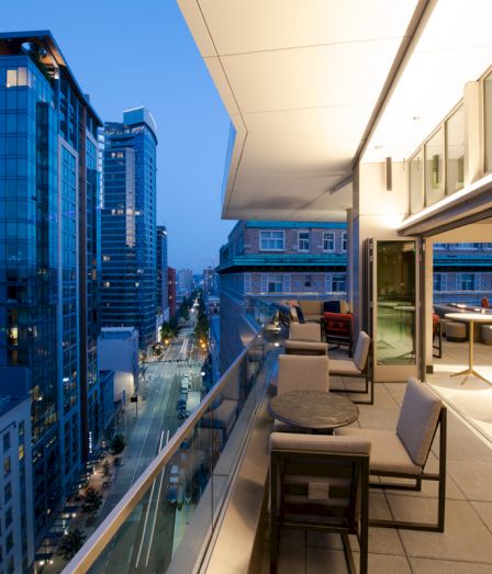 Modern high-rise balcony with patio seating overlooks a busy city street between tall buildings at dusk.