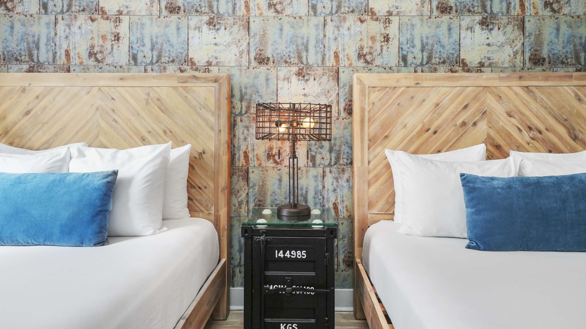 Two neatly made twin beds with wooden headboards, white linens, blue pillows, and a shared vintage nightstand between them.