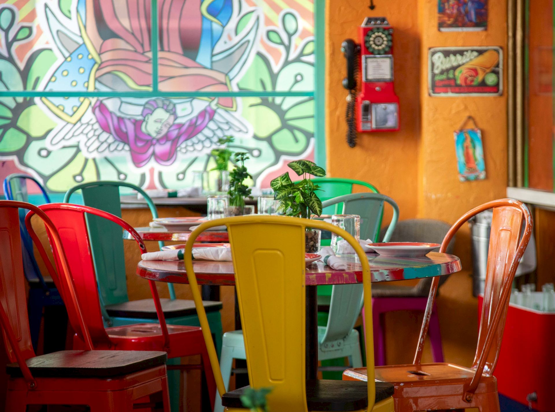 A colorful caf&eacute; patio with bright chairs, a table set with plants and glassware, and a graffiti wall in the background.