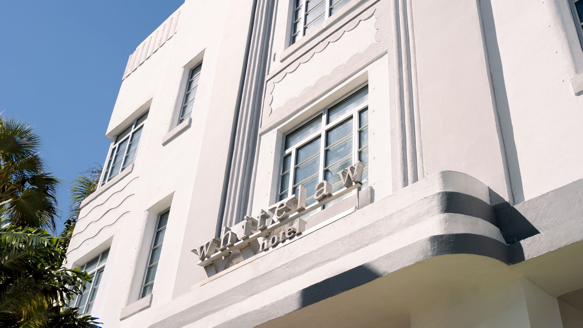An art deco white building with rounded corners and palm trees, sign reads &ldquo;Windelay &hellip;&rdquo; or &ldquo;Lown a&hellip;&rdquo; on the fa&ccedil;ade, sunny day.