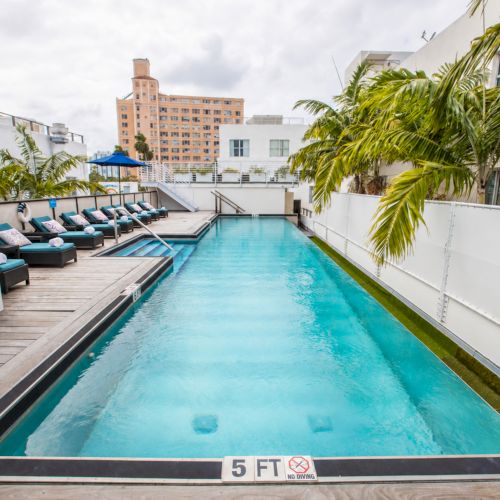 A rooftop courtyard pool with blue water, lounge chairs, palm trees, and surrounding apartment buildings under an overcast sky.
