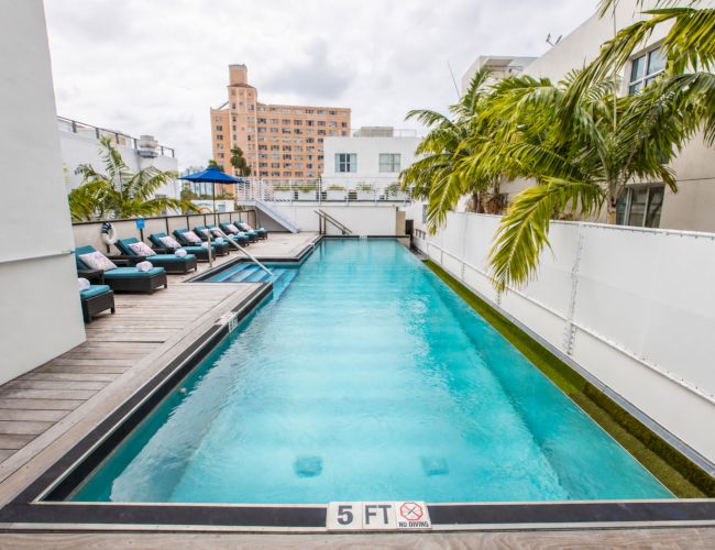 A rooftop courtyard pool with blue water, lounge chairs, palm trees, and surrounding apartment buildings under an overcast sky.