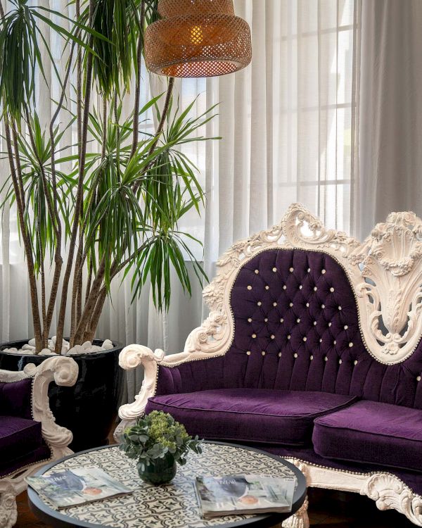 Elegant living room with a white ornate sofa, purple tufted cushions, a glass coffee table, lush plants, and warm woven pendant lighting.