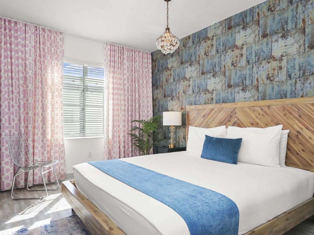 A bright hotel-style bedroom with a wooden platform bed, blue accent runner and pillow, blue textured wall, pink curtains, and a chandelier.