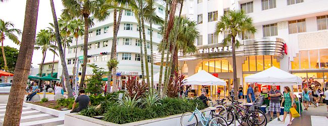 A sunny campus scene with palm trees, bikes, and people gathering outside a white modern building with umbrellas and storefronts, lively and tropical.