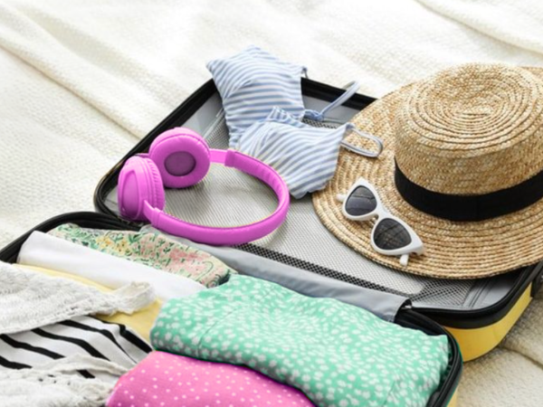 A packed suitcase with clothes, hat, sunglasses, pink headphones, and pastel swimwear ready for a sunny getaway.