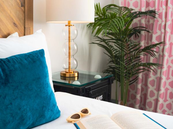 A cozy bedroom scene with a bed featuring teal pillows, a bedside table, a lamp, sunglasses, an open notebook, and a plant by pink patterned curtains.