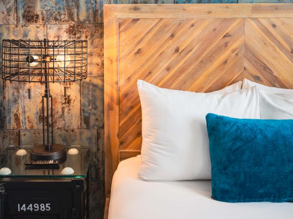 A rustic bedroom scene with a wooden chevron headboard, white bedding, a teal throw pillow, and a vintage-style telephone table with a metal lamp against a weathered wall.