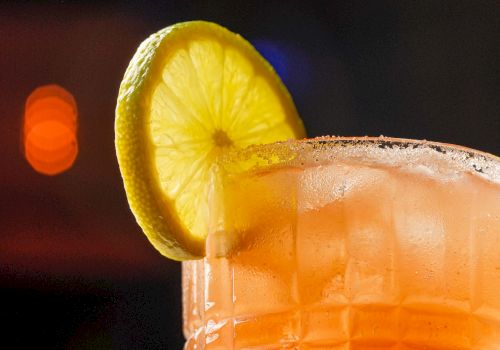 A refreshing orange-pink cocktail in a textured glass with a lemon wheel garnish on the rim, set against a dim, blurred background. It's a vibrant, upscale drink.