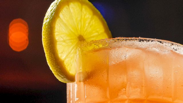 A refreshing orange-pink cocktail in a textured glass with a lemon wheel garnish on the rim, set against a dim, blurred background. It's a vibrant, upscale drink.