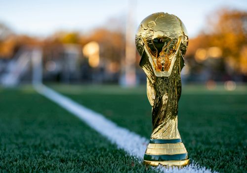 A golden FIFA World Cup trophy sits on a green football pitch near the white line, with a blurred stadium background.
