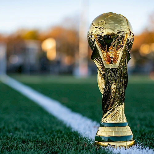 A golden FIFA World Cup trophy sits on a green football pitch near the white line, with a blurred stadium background.