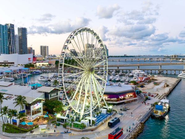 A coastal cityscape with a large Ferris wheel by the waterfront, modern high-rise buildings, boats, and a bustling pier at dusk.