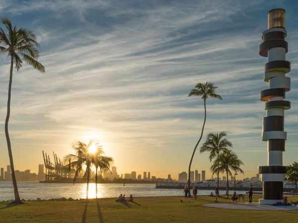 A sunny beach scene with a few tall palm trees, a striped spiral tower, and people enjoying the shore as the sun sets over the water.
