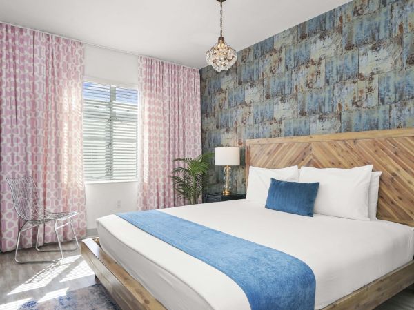 A modern bedroom with a wooden platform bed, blue accent throw and pillow, pink patterned curtains, a textured blue wall, and a small chandelier.
