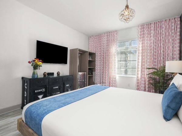 A bright, modern bedroom with a white and blue color scheme, a king bed, a TV stand, pink patterned curtains, and decorative plants.