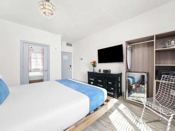 A bright hotel-style bedroom with a white and blue theme, a wall-mounted TV, open wardrobe, mirrored dresser, and sunlit seating, stylishly simple.