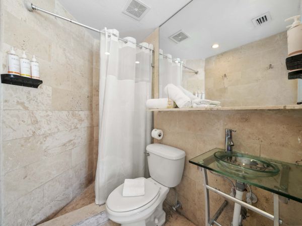 A compact bathroom with a shower curtain, toilet, glass sink on a metal frame, shelf with toiletries, and a mirror area above.