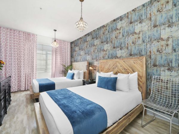 A modern hotel room with two queen beds, blue runners and pillows, patterned blue feature wall, pink curtains, a small desk, and a metal chair. End.