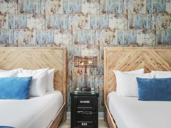 Two neatly made twin beds with white sheets and blue pillows, a shared nightstand with a rustic lamp, and distressed patterned walls.