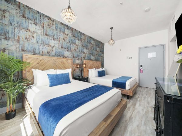 A bright hotel room with two wooden beds, blue accents, a painted accent wall, potted plant, and a desk area with fresh flowers.