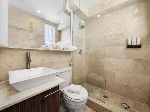 A modern bathroom with a rectangular vessel sink, wooden vanity, wall mirror, toilet, and a glass-enclosed shower with beige tile and a small shelf of toiletries.
