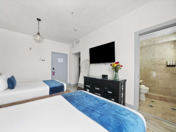 A bright hotel room with two beds, blue accents, a wall-mounted TV, a dresser with flowers, and an open ensuite bathroom.