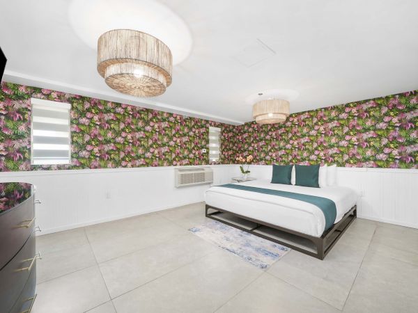 A bright, modern bedroom with bold floral wallpaper, a white platform bed with teal accents, floating desk area, and soft pendant lights.