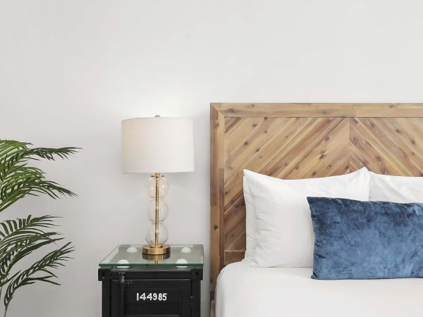 A minimalist bedroom with a wooden headboard, white bedding, a blue throw pillow, a glass-topped nightstand, a brass lamp, and a potted plant beside the bed.