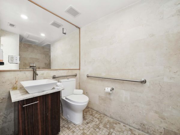 A modern bathroom with a single-sink vanity, wall mirror, toilet, grab bar, light fixtures, and beige tiled walls and floor, compact and accessible.