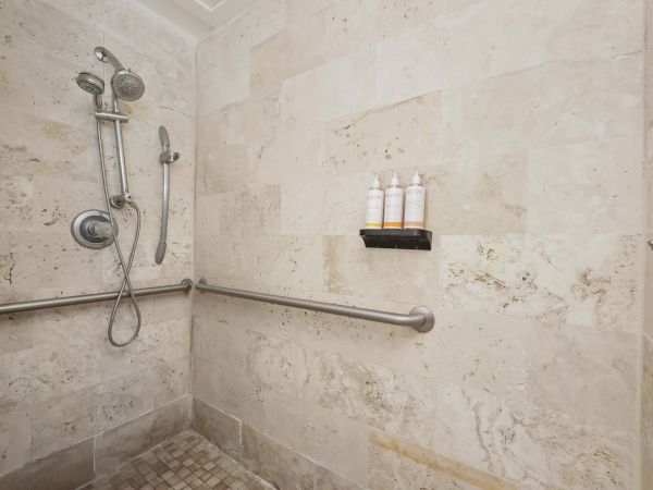 A walk-in shower with tiled walls, a handheld shower, two fixed heads, a grab bar, and a small shelf holding three bottles.