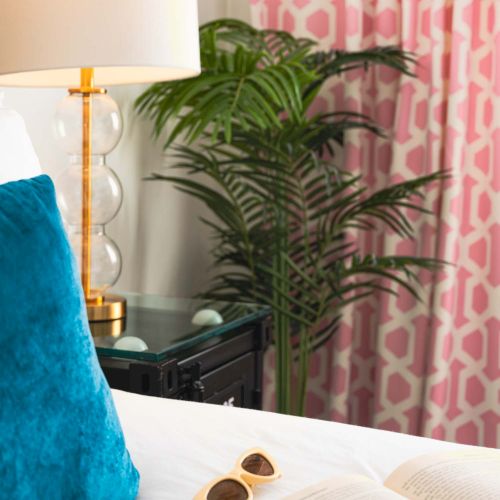 A cozy bedside scene with a blue velvet pillow, open book, sunglasses, lamp, and pink patterned curtains beside a lush houseplant.