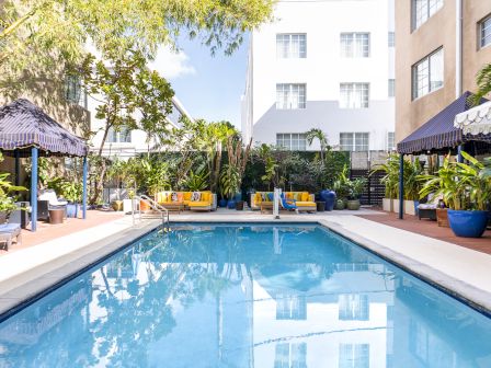 A resort-style rectangular outdoor pool surrounded by lounge chairs, shaded cabanas, tropical plants, and a white building backdrop.