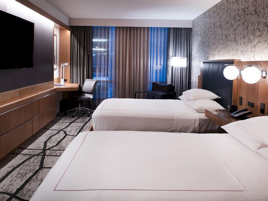 A modern hotel room with two white beds, dark wood panels, and wall-mounted TVs.