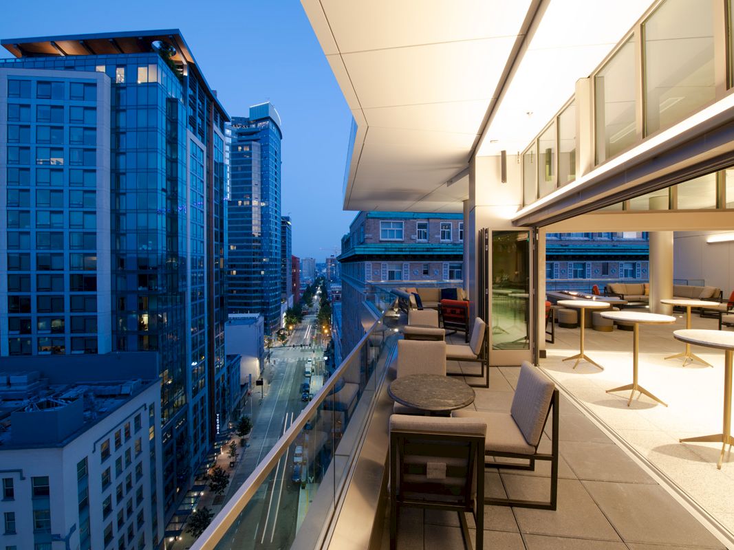 A modern high-rise rooftop terrace with lounge seating overlooks a city street and skyline at dusk.